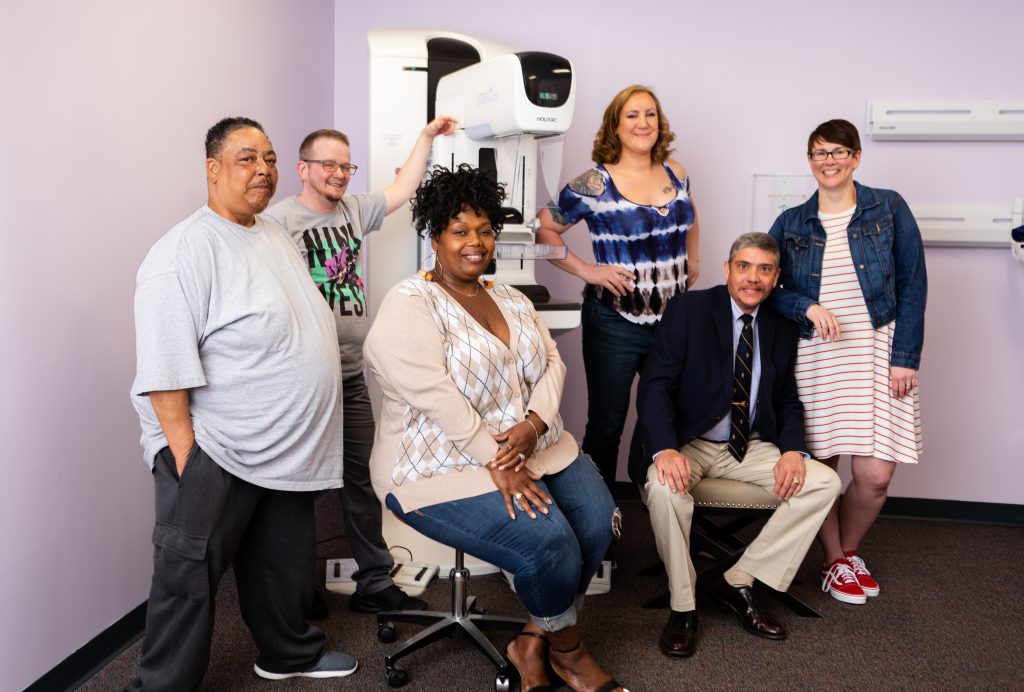 People of various genders and races standing in a mammogram exam room.